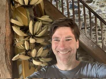 A smiling man takes a selfie indoors near a staircase. Behind him hangs a large cluster of bananas attached to a thick tree stem, creating a tropical vibe.