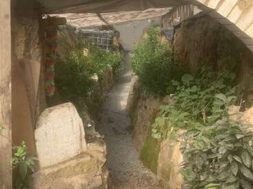 This image shows a narrow, dirt-walled trench lined with green plants. There's a gravel path, a stone tablet, and a wooden shelter overhead, creating a rustic atmosphere.