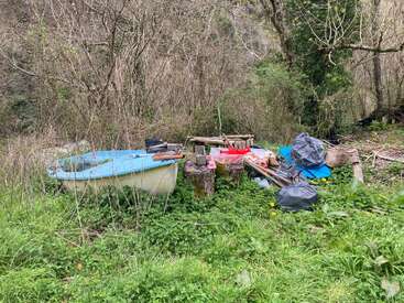 Un pequeño barco azul y blanco yace abandonado entre la maleza, rodeado de bolsas de basura tiradas, tablones de madera, recipientes de plástico y restos diversos en plena naturaleza.