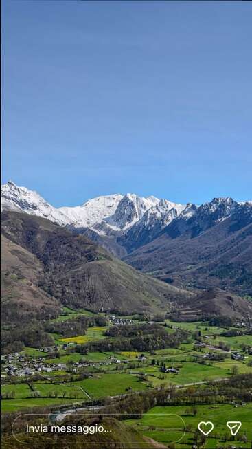 Un paisaje pintoresco de un valle verde salpicado de casas, que se extiende hasta colinas boscosas y imponentes montañas nevadas bajo un cielo azul y despejado.