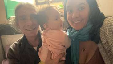 Un hombre, una mujer y un bebé sonrientes posan muy juntos en el interior de una casa. El bebé va vestido de rosa y la mujer lleva un pañuelo turquesa, lo que crea un momento familiar cálido y feliz.