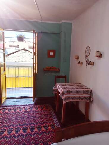 La lumière du soleil pénètre dans une pièce confortable avec un tapis à motifs, une petite table et un attrape-rêves accroché au mur. Des portes ouvertes mènent à un balcon ensoleillé à l'extérieur.