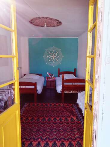 Cette chambre confortable dispose de deux lits simples, d'un mur d'accent turquoise avec un motif de mandala, d'un tapis à motifs vibrants, de portes jaunes et d'une douce lumière naturelle.
