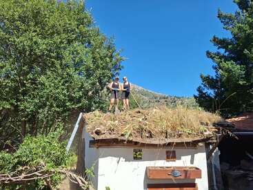 Dos personas en una azotea verde y cubierta de hierba, rodeadas de frondosos árboles y un cielo azul despejado, con una montaña lejana al fondo. Día luminoso y soleado.