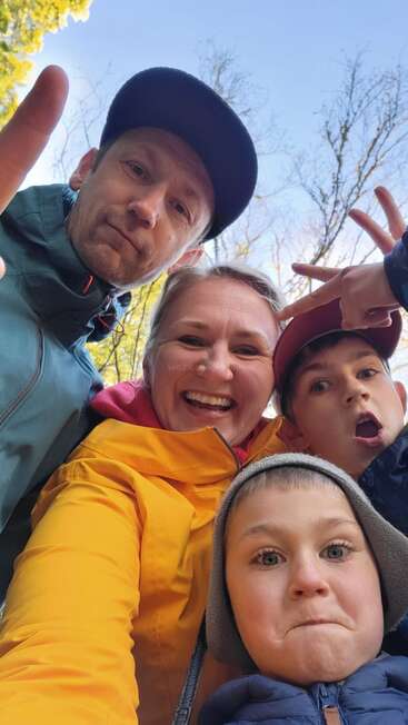 Una alegre selfie familiar al aire libre, en la que aparecen dos adultos y dos niños. Van bien abrigados con chaquetas y gorros, y sonríen y ponen caras graciosas bajo un cielo radiante.