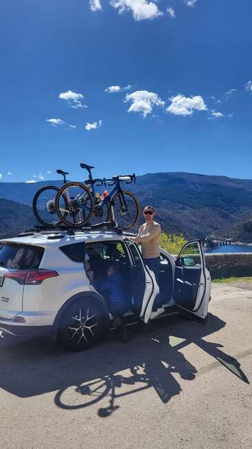 Un hombre está junto a un todoterreno blanco con dos bicicletas en el techo. Bajo un cielo azul brillante se divisan unas pintorescas montañas, un río y un puente.