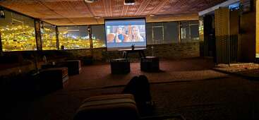 Eine gemütliche Dachterrassen-Lounge mit gemusterten Teppichen und Kissen, auf denen ein Projektor einen Film abspielt. Die Lichter der Stadt scheinen hell durch die großen Fenster und schaffen eine warme Atmosphäre.