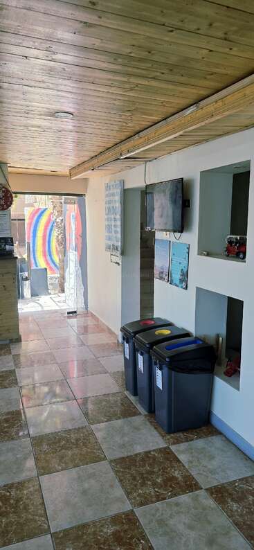 Ein heller Flur mit gefliestem Boden, Holzdecke, drei Wertstofftonnen, Wandnischen, Postern, TV-Bildschirm und einem bunten Regenbogen-Wandbild, das durch die Glastür zu sehen ist.