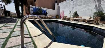 Dieses Bild zeigt einen kleinen Swimmingpool im Freien, umgeben von gefliesten Böden, hölzernen Sitzgelegenheiten, Palmen, Wandgraffiti und hellem Tageslicht, das Schatten wirft.