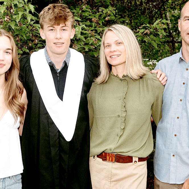 Ein junger Mann in einer Abschlussrobe steht mit seiner Familie - zwei Frauen und einem Mann - draußen, lächelt und posiert gemeinsam vor grünen, belaubten Büschen.