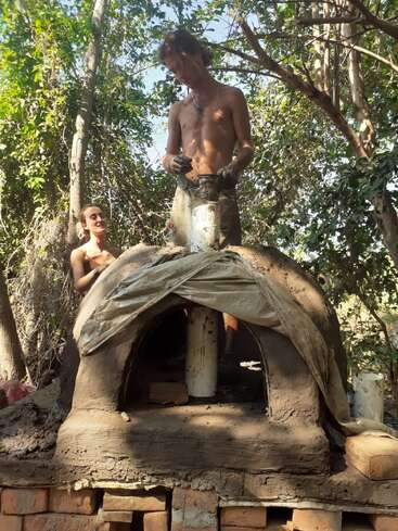Dos personas, con el torso desnudo, están construyendo un horno de barro al aire libre en una zona boscosa. Una de ellas está moldeando el barro en la parte superior, mientras que la otra observa sonriendo.