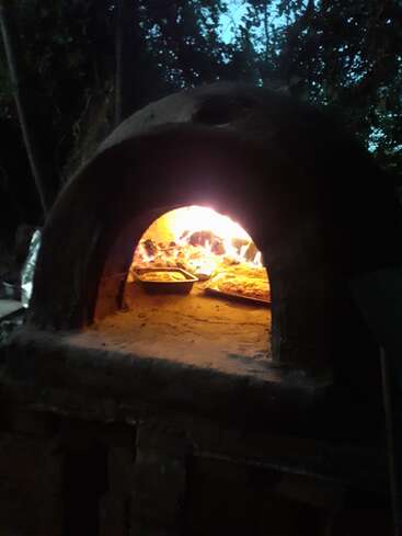 Un horno de leña rústico al aire libre brilla cálidamente al atardecer, con bandejas de comida horneándose en su interior. Rodeada de árboles, la escena resulta acogedora y tentadora.