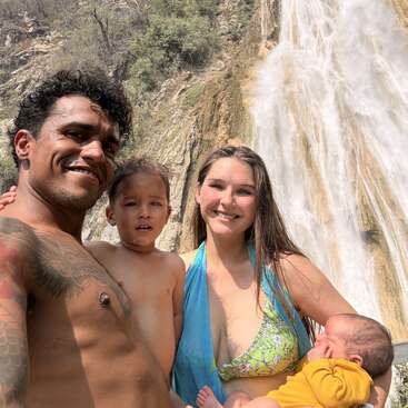 Une joyeuse famille de quatre personnes pose pour une photo devant une magnifique cascade. Les parents sourient, serrant leurs deux jeunes enfants contre eux.