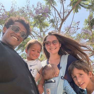 A smiling family of five poses outdoors under a sunny sky with blooming trees. The parents wear sunglasses; the baby is in a front carrier. Joyful moment.