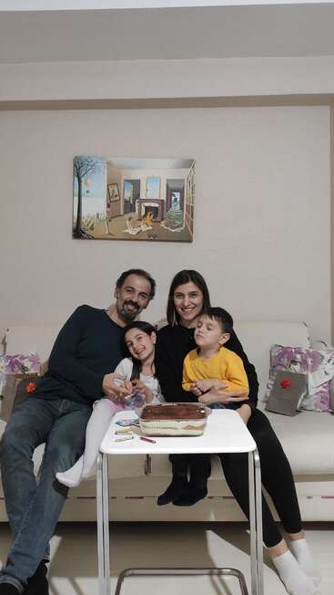 A happy family of four sits on a couch, smiling, with a cake on a table in front of them. Colorful art decorates the wall.