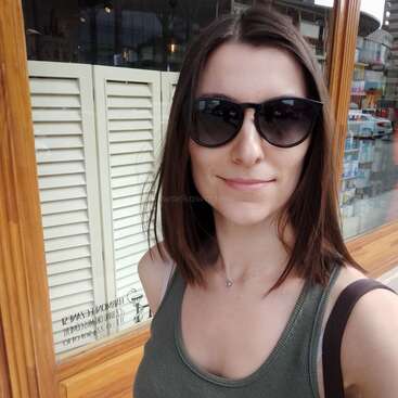 A woman with straight brown hair and sunglasses smiles outdoors. She wears a green tank top and carries a bag, standing near a window with wooden frame.