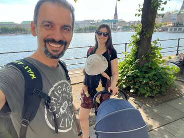 A smiling man takes a selfie by a waterfront with a woman, baby in a carrier, stroller, green trees, and city buildings in the background.