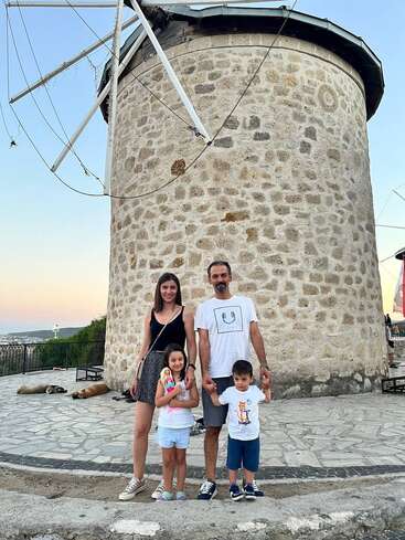 A smiling family of four poses in front of an old stone windmill at sunset. Two dogs rest on the ground nearby, creating a peaceful scene.