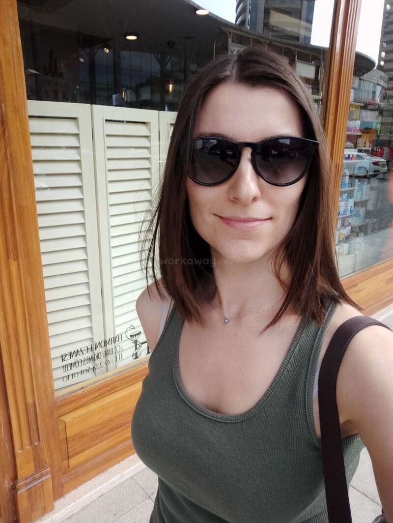 A woman with straight brown hair and sunglasses smiles outdoors. She wears a green tank top and carries a bag, standing near a window with wooden frame.