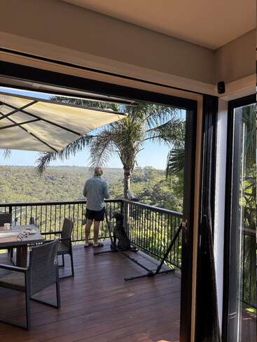 Une personne et un chien se tiennent sur un balcon en bois, face à une vallée verdoyante. Une porte ouverte, du mobilier d'extérieur et des palmiers viennent compléter cette scène paisible.