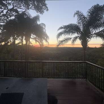A serene sunset view from a wooden deck, overlooking lush green forest and tall palm trees, with a tranquil sky painted in soft evening hues.