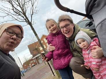 Une famille joyeuse pose en plein air. Une femme porte un hijab et des lunettes, un homme à lunettes serre dans ses bras deux jeunes filles vêtues de vestes d'hiver, tous souriant joyeusement ensemble.