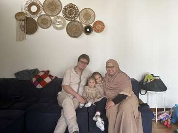 Une famille de quatre personnes est assise ensemble sur un canapé bleu foncé, souriante. Une décoration murale tissée est accrochée au-dessus d'eux. Une scène familiale chaleureuse, joyeuse et accueillante.