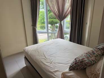 This cozy bedroom features a neatly made bed, pastel pillowcases, large windows with sheer curtains, and a view of green trees and parked cars outside.