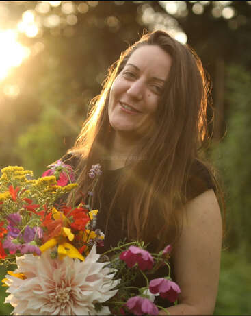 Une femme aux longs cheveux bruns sourit chaleureusement, tenant un bouquet de fleurs coloré. La lumière dorée du soleil brille derrière elle, créant une atmosphère douce, lumineuse et naturelle en plein air.