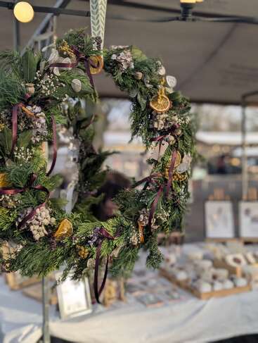 Une couronne festive décorée de tranches d'orange, de rubans et de pommes de pin est suspendue à un étal de marché. L'arrière-plan présente des objets artisanaux et des produits en céramique sur des tables.
