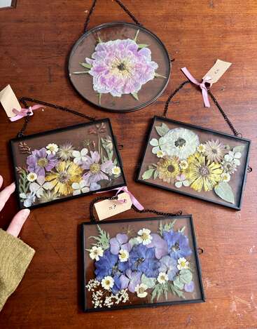 Quatre cadres en verre avec des bordures noires présentent des fleurs pressées et colorées. Trois sont rectangulaires, un est circulaire. Tous les cadres sont munis d'une chaîne de suspension et d'une étiquette.