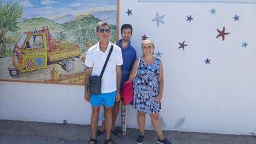 Três pessoas posam sorridentes em frente a uma parede branca decorada com estrelas-do-mar e um mural colorido de um tuk-tuk em um cenário rural.