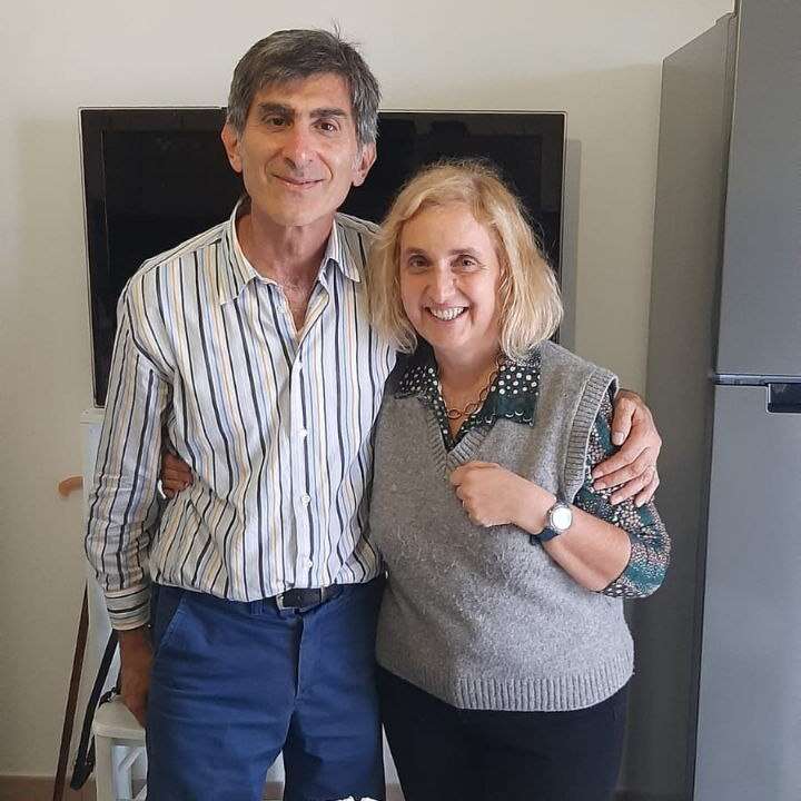 Um homem e uma mulher sorriem juntos em um ambiente interno. O homem usa camisa listrada e calça azul. A mulher usa um colete cinza e uma blusa estampada. Atmosfera calorosa.