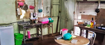 This is a modest, rustic kitchen with basic utensils, a sink, a round table, dish rack, washing machine, and metal mesh window panels for ventilation.