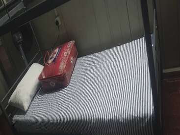 A neatly made single bunk bed with striped bedsheet, a white pillow, and a red bag placed at the head of the bed. Room appears small.