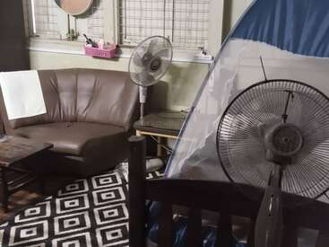 A cozy living space features a brown sofa, two standing fans, a geometric patterned rug, a small table, a tent, and windows with bars.