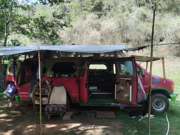 Un camping-car rouge et vert est garé en plein air ; il est équipé d'un auvent de fortune, de mobilier d'extérieur et de matériel de camping, et se trouve au milieu de la nature, entouré d'arbres sur un terrain herbeux.
