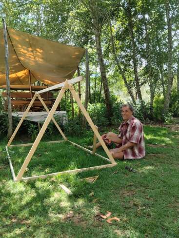 Une personne est assise sur l'herbe et construit un cadre en bois à l'aide d'outils. Derrière elle se trouve un abri recouvert d'une bâche, entouré d'arbres verdoyants et d'une nature luxuriante.