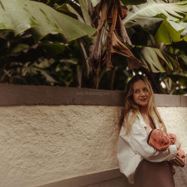 A woman with long hair, wearing a white shirt, leans against a textured wall while holding a baby. Lush, green tropical plants hang overhead, creating a serene atmosphere.