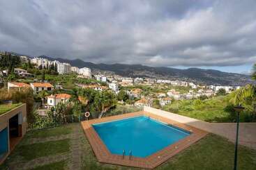 A blue swimming pool overlooks a lush hillside dotted with houses and buildings. Mountains rise in the background under cloudy skies, creating a scenic, tranquil view.