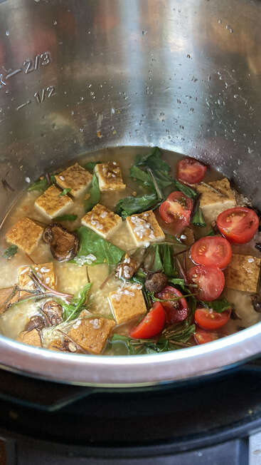 Inside a metal pot, tofu cubes, cherry tomatoes, mushrooms, and leafy greens simmer in broth, creating a healthy, colorful, plant-based soup or stew.