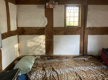 A cozy room with wooden beams and white plaster walls. Small window, bed with leopard-print blanket and green pillow, minimal decor, suitcase on the side.