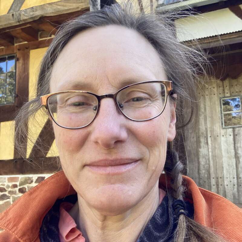 A woman with glasses and a braid smiles gently at the camera, wearing an orange jacket, standing in front of a rustic wooden and stone building.
