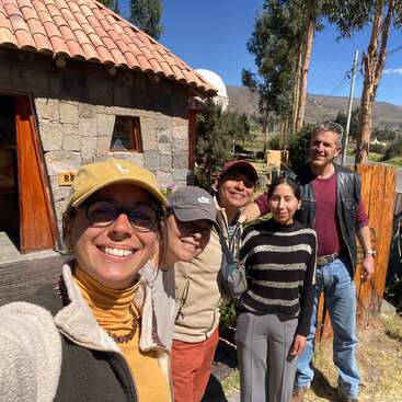 Five people smile outdoors in front of a rustic stone building with a tiled roof, surrounded by tall trees and mountains under a clear blue sky.
