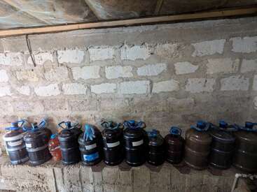 Several large plastic jugs, mostly dark in color, are lined up on a shelf against an unfinished stone wall. One pair of gloves sits atop them.