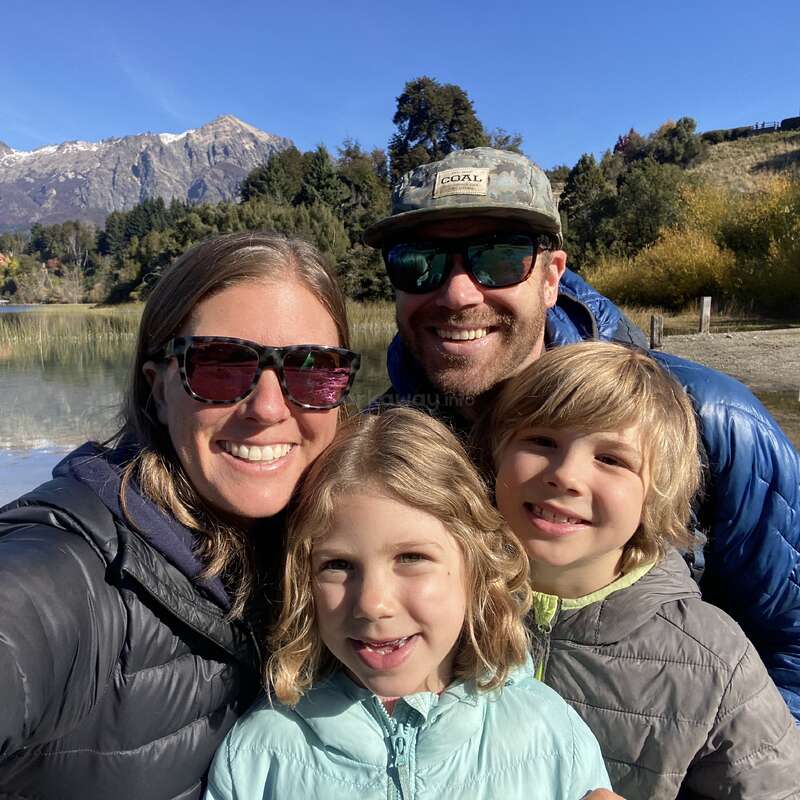 Uma família feliz de quatro pessoas tira uma selfie ao ar livre à beira de um lago, cercada por árvores e montanhas, todos sorridentes e vestindo jaquetas e óculos escuros.