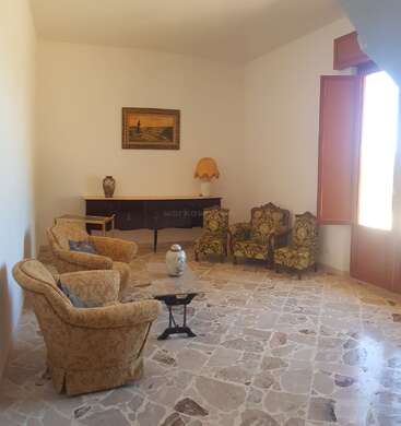 A spacious, bright living room with vintage armchairs, a marble floor, a sideboard holding a lamp and vase, a framed painting, and an open window.