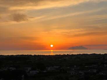 A beautiful sunset over the sea, with golden and orange hues in the sky, calm waters reflecting the sun, and distant mountains silhouetted on the horizon.
