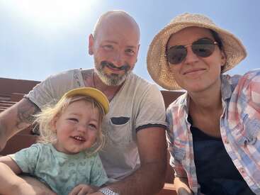 A cheerful family of three poses outside in the sunlight. The child wears a yellow cap, the man has a beard, and the woman wears sunglasses and a sunhat.