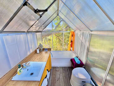 This image shows a greenhouse-style bathroom with a sink, wooden countertop, bathtub, stool, and towels, featuring transparent walls for a bright, nature-inspired bathing experience.